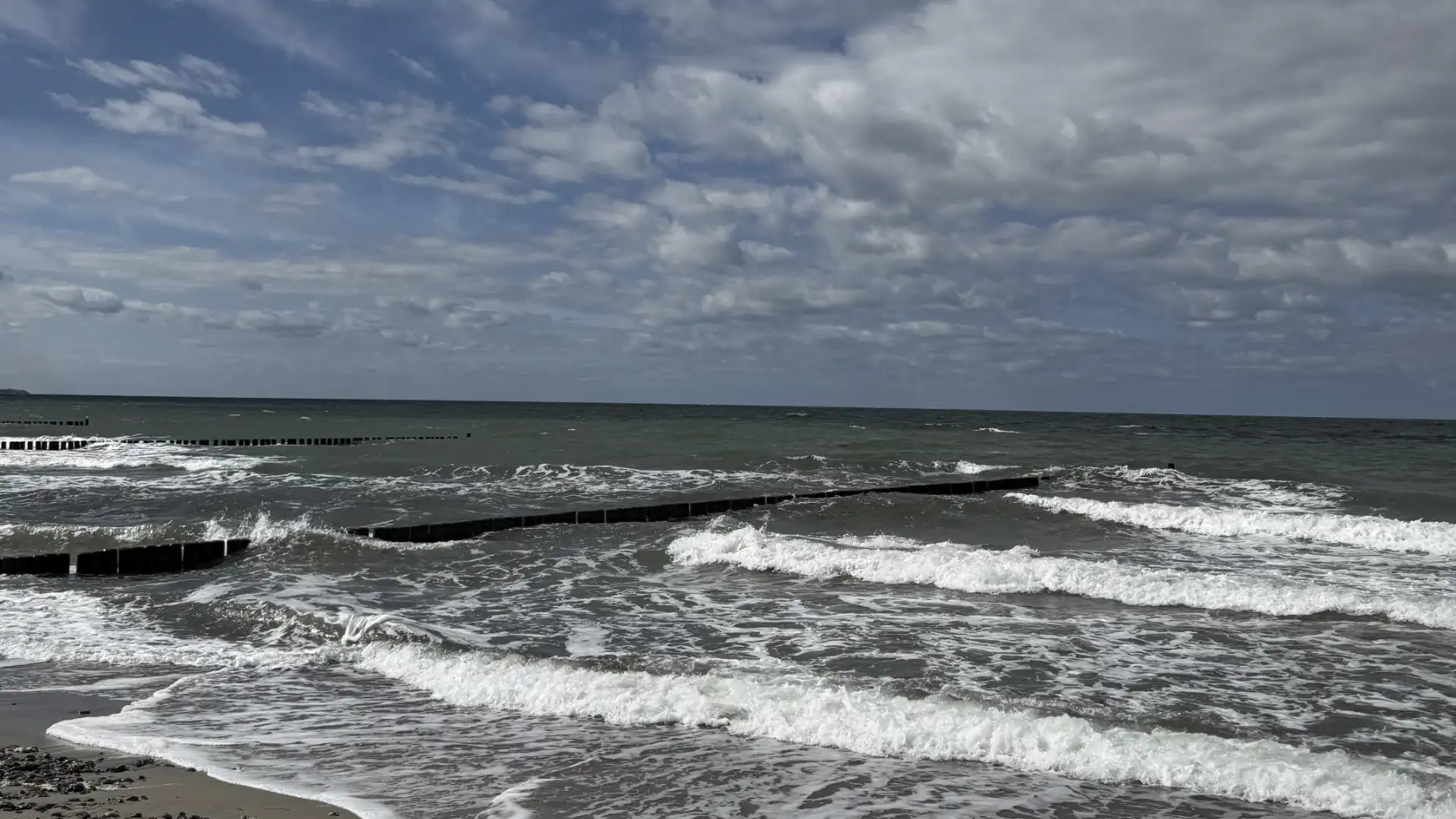Titelbild zum Artikel „Ferienzeit – Zeit zum Nachdenken“. Zu sehen ist ein bewölkter Himmel über einer See, die zum Ufer hin mit weiß-schäumenden Wellen heranrollt. In das bewegte Gewässer ragen von links her hölzerne Stege.